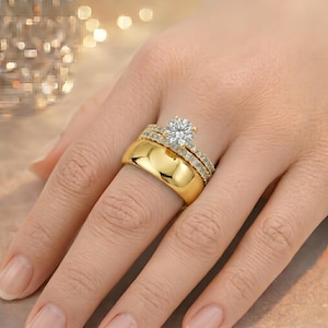 Könnte beinhalten: Ein goldener Ehering-Satz. Das Set besteht aus einem breiten Band und einem diamantbesetzten Band mit einem großen, rund geschliffenen Diamanten als Mittelstein. Die Ringe befinden sich an einem Finger und zeigen ihre polierten, reflektierenden Oberflächen.