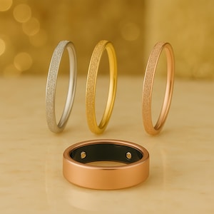 May include: A collection of rings in silver, gold, and rose gold, with a textured surface. A larger, rose gold ring with a black interior is in the foreground. The rings are displayed on a light-colored surface.
