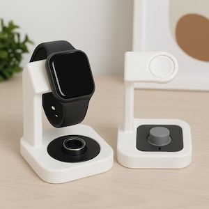 May include: Two white watch charging stands. One holds a black smartwatch with a black strap. The other stand is empty, with a grey button on the base. Both stands have a minimalist design.