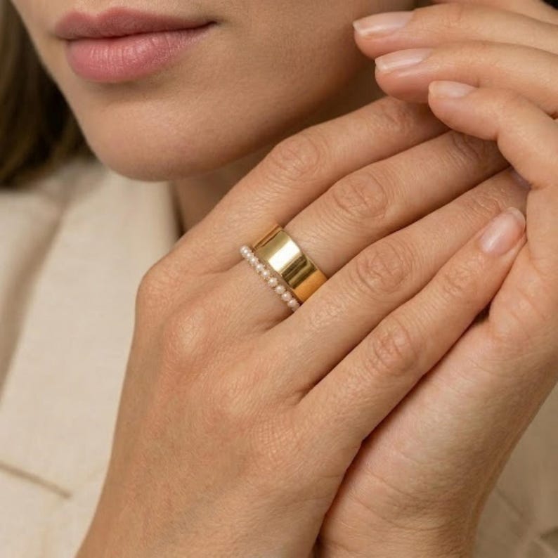 May include: A gold ring with a wide band and a row of small pearls. The ring is on a person's finger. The person is wearing a cream-colored blazer.