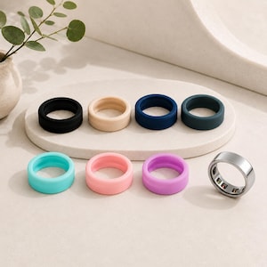Peut inclure: Une collection de sept bagues en silicone noir, beige, bleu marine, bleu sarcelle, turquoise, rose clair et lavande, disposées à côté d'une bague connectée en métal argenté avec des capteurs internes visibles sur la bande intérieure.