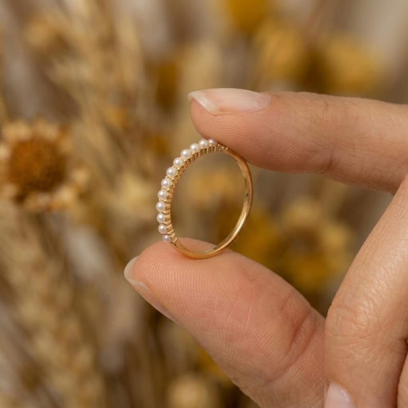 May include: A gold ring with a row of small, white pearls. The ring is held between two fingers, showcasing its delicate design. The background is blurred, with warm tones.