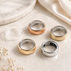 Puede incluir: Una colección de cuatro anillos lisos de colores sólidos en tonos oro rosa, oro amarillo y plata, dispuestos junto a un anillo transparente. Cada anillo presenta un diseño de banda simple y redondeado con un acabado metálico pulido y brillante.