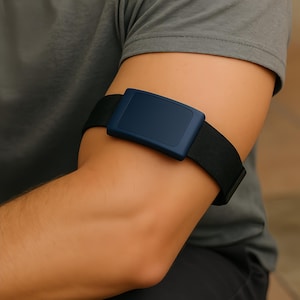 Può includere: Un cinturino nero con un dispositivo rettangolare blu navy è avvolto attorno al braccio di una persona. Il dispositivo è centrato sul braccio e sembra essere in plastica. La persona indossa una maglietta grigia.