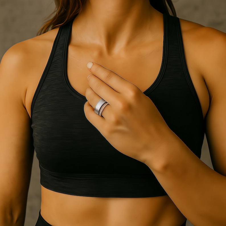 Puede incluir: Un anillo plateado con la parte superior plana y una banda texturizada se usa en un dedo. El anillo se combina con un sujetador deportivo negro. El fondo es gris neutro.