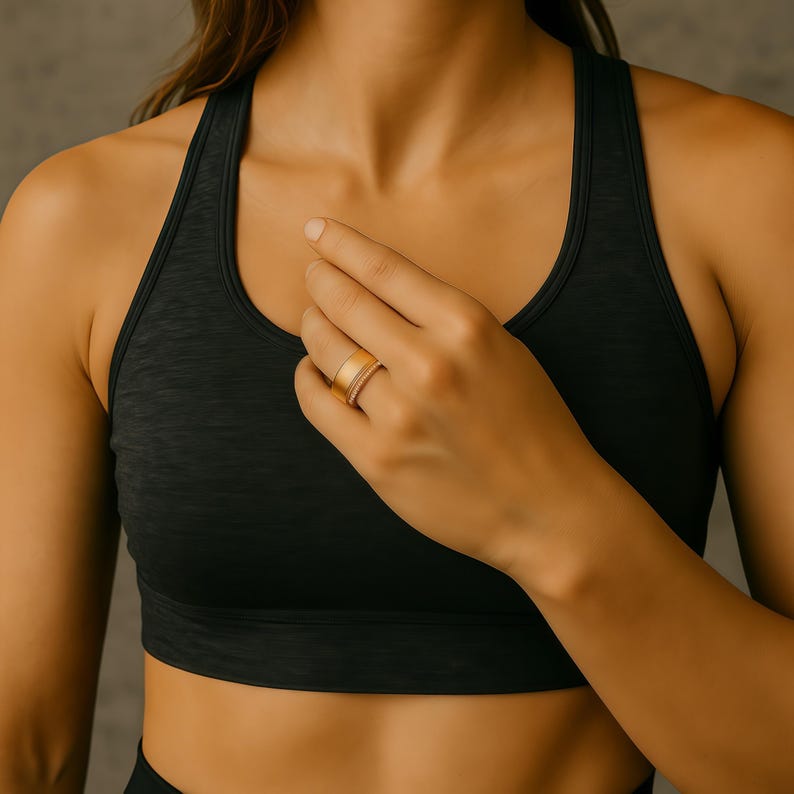 Puede incluir: Un anillo dorado con una banda ancha y una banda m&aacute;s delgada, posiblemente con diamantes. El anillo se lleva en un dedo. La persona lleva un sujetador deportivo negro.