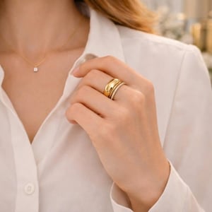 Può includere: Una collana d'oro con un piccolo pendente quadrato e due anelli d'oro su un dito. Un anello è una fascia spessa, l'altro è una fascia più sottile con piccole pietre scintillanti. La persona indossa una camicia bianca abbottonata.