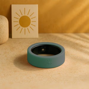 May include: A teal smart ring with a black interior and a small sun icon on the side. The ring is on a light brown surface, with a decorative square tile featuring a sun design in the background.