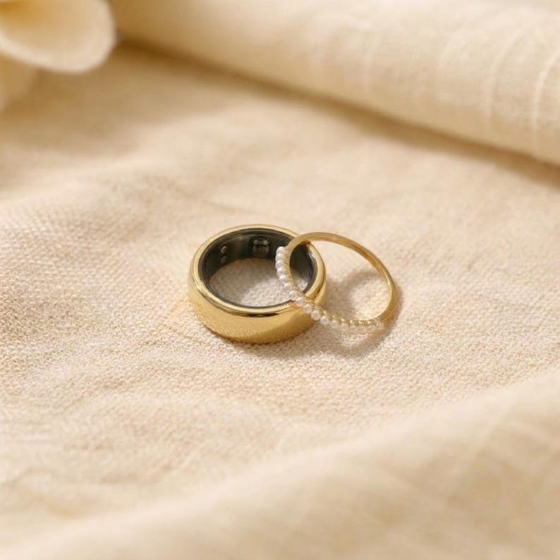 May include: A gold smart ring and a delicate gold ring with small white pearls. The smart ring is thick and has a dark interior. Both rings are on a beige fabric, creating a soft, neutral background.