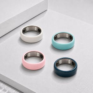 Op de afbeelding: Vier smart rings gerangschikt in een vierkant. Elke ring heeft een gladde, effen gekleurde buitenband in wit, lichtblauw, zachtroze en donker marineblauw, met een metalen binnenvoering die zichtbare elektronische sensoren en componenten bevat.
