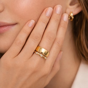 May include: A close-up of a hand wearing two gold rings. The top ring is a wide band, and the bottom ring has a delicate design with three small, clear stones. A gold hoop earring is visible in the background.