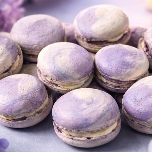 May include: A close-up of several lavender-colored macarons with a marbled effect. The cookies have a creamy filling and are arranged in a cluster. The macarons are a light purple color with a white swirl.