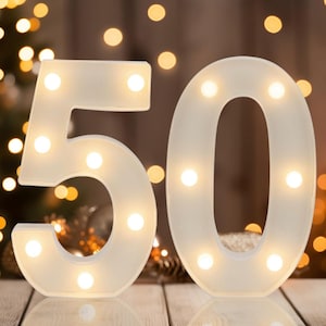 May include: White illuminated number 50 with warm white lights. The numbers are on a wooden surface with blurred background lights. The lights are evenly spaced within the numbers, creating a soft glow. Perfect for a birthday or anniversary.