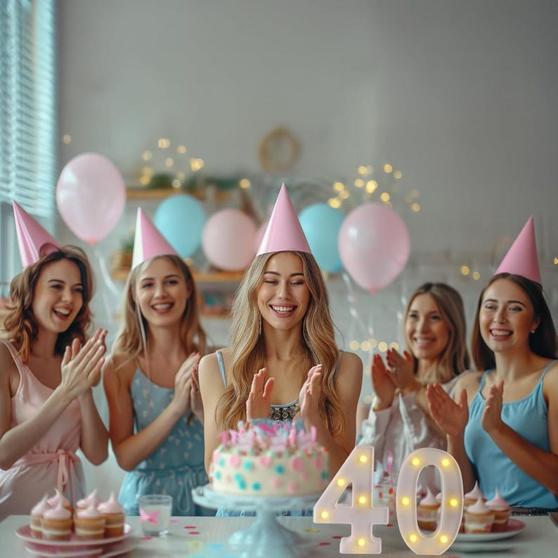 May include: A group of women celebrating a birthday. A cake with candles sits on a table, with the number "40" lit up beside it. Pink and blue balloons and party hats add to the festive atmosphere.