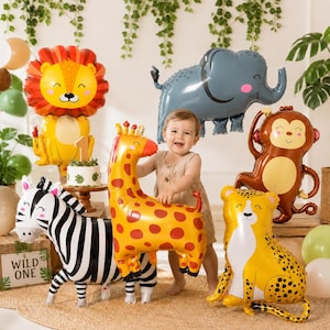 May include: A collection of animal-shaped foil balloons including a lion, elephant, monkey, giraffe, zebra, and cheetah. The balloons feature cartoon-style faces and vibrant colors. A small cake with a number one topper sits on a wooden stand.
