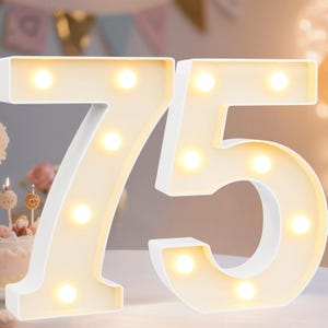 May include: White, illuminated numbers '75' with warm white lights. The numbers are likely for a birthday or anniversary celebration. The background includes a cake and party decorations.