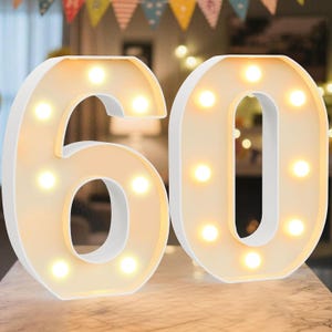 May include: White illuminated number 60, ideal for a birthday or anniversary. The numbers are lit with warm white lights, creating a soft glow. The numbers are made of a white material and are placed on a light-coloured surface.