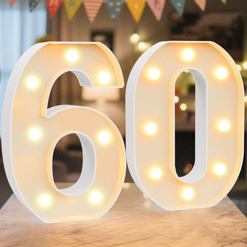May include: White illuminated number 60, ideal for a birthday or anniversary. The numbers are lit with warm white lights, creating a soft glow. The numbers are made of a white material and are placed on a light-coloured surface.