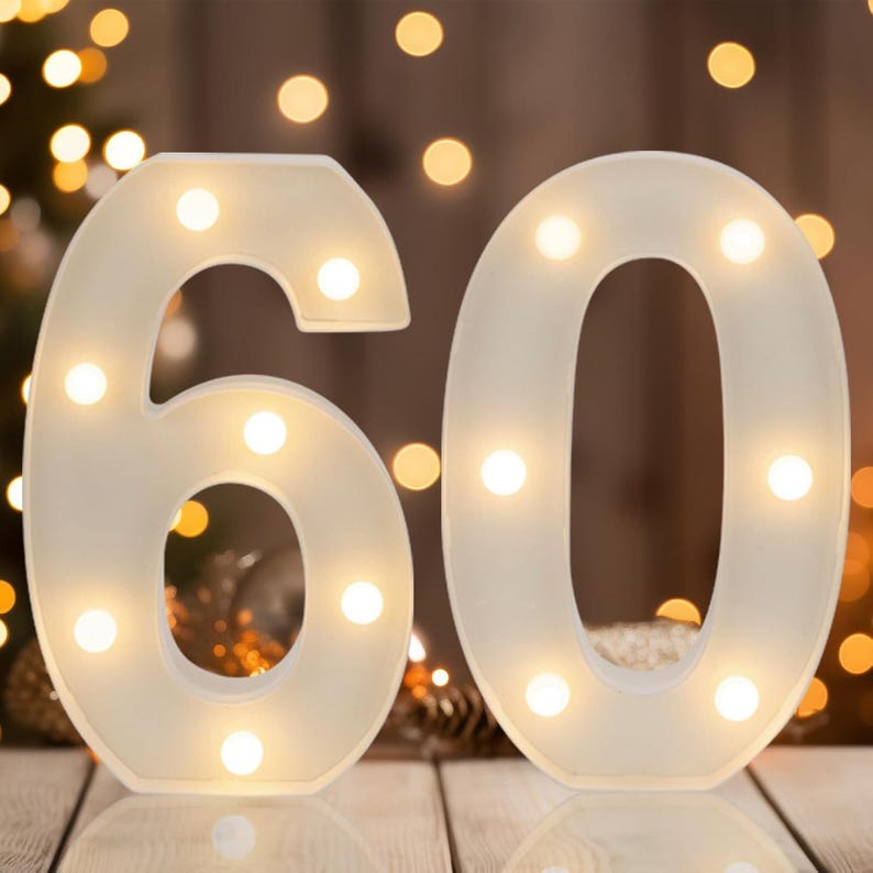 May include: A pair of large, white, three-dimensional number signs representing 60, each featuring integrated warm white LED lights embedded within the structure to create a glowing effect across the surface of the numbers.