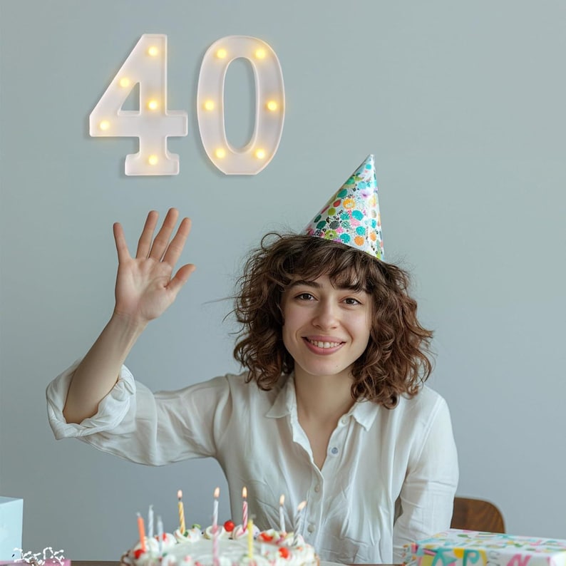 May include: A woman wearing a party hat smiles at the camera, with a birthday cake in front of her. Above her, a white illuminated number 40 is mounted on the wall. The cake has lit candles.