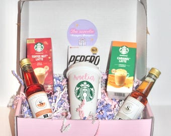 Puede incluir: Una caja de regalo rosa llena de una taza de café, dos cajas de café Starbucks y dos pequeñas botellas de jarabe aromatizado. La taza es blanca con un logotipo de Starbucks verde y el nombre "Amelia" escrito en ella. Las cajas de café son para Toffee Nut Latte y Caramel Latte. La caja de regalo tiene una pegatina que dice "The sweetie" en ella.
