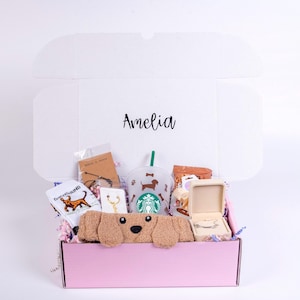 May include: A pink gift box filled with dog-themed items, including a plush dog, socks, a Starbucks cup, and jewelry. The box is open, revealing the contents, and the lid is inscribed with the name "Amelia".