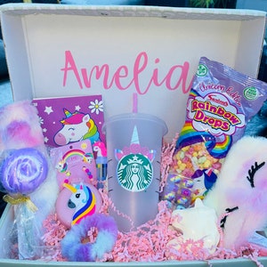 May include: A pink gift box with the name Amelia on the lid. The box is filled with unicorn-themed gifts, including a notebook, a unicorn-shaped lollipop, a fluffy unicorn tail, a unicorn-themed reusable cup, a unicorn-shaped soap, and a bag of rainbow-coloured candy.
