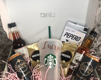Puede incluir: Una caja de regalo llena de artículos con temática de café, que incluyen un vaso Starbucks personalizado, cápsulas de café y bebidas de café enlatadas. La caja está forrada con papel de seda rosa e incluye una pequeña bolsa de palitos de chocolate Pepero.