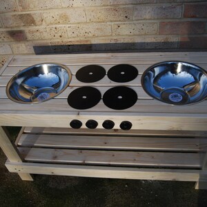 May include: A wooden play kitchen with two stainless steel bowls, four black hob covers, and four black knobs. The play kitchen is made of unfinished wood and has a lower shelf.