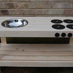 May include: A wooden play kitchen with a stainless steel sink and four black burner circles. The play kitchen also has four black knobs and a lower shelf for storage. The wooden structure is made of light-coloured wood.