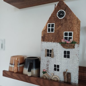 Könnte beinhalten: Eine Holzhausfigur mit weiß gestrichenem Äußeren und braunem Dach. Das Haus hat Fenster, eine Tür und ein kleines Fahrrad davor. Die Figur steht auf einem Holzregal mit zwei Bechern.
