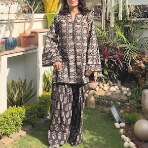 Op de afbeelding: Een zwart-witte tuniek met lange mouwen, een herhalend bloemenpatroon en een split-halslijn, gecombineerd met een bijpassende wijde broek met een vergelijkbare bloemenprint. De outfit wordt afgemaakt met zwarte slippers en een zonnebril.