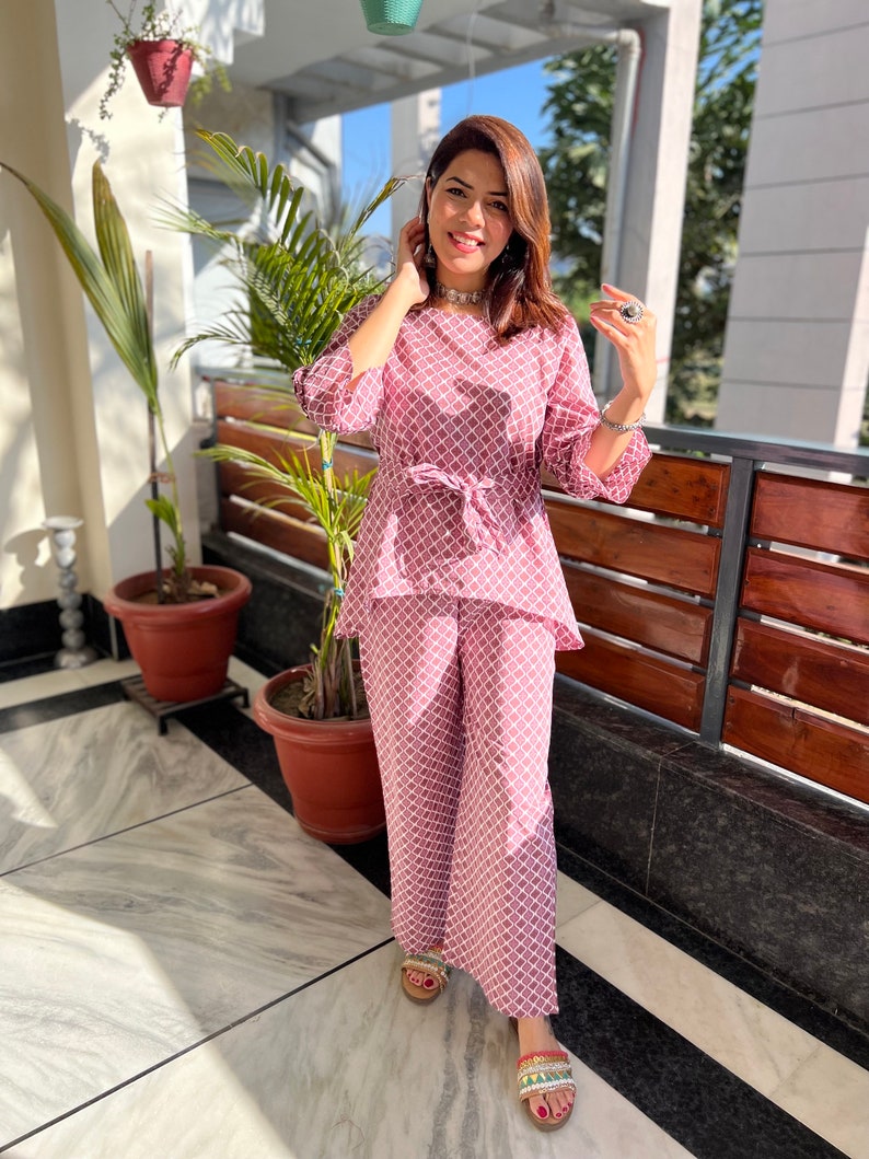 May include: A pink and white patterned pantsuit with a tie waist. The pantsuit is made of a lightweight fabric and has a relaxed fit.