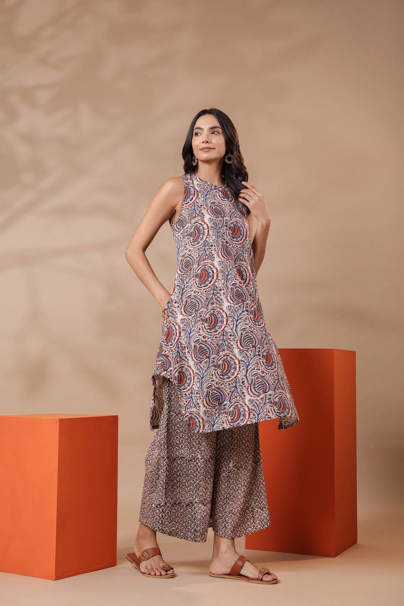 May include: A sleeveless tunic and wide-leg pants set. The tunic features a cream-colored base with a paisley pattern in red and blue. The pants have a complementary brown and cream pattern. The model is wearing brown sandals.