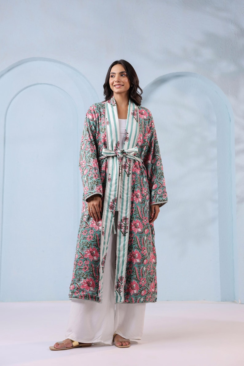 May include: A long, floral-print robe in shades of teal and pink, with a striped accent on the lapel and belt. The robe is worn with white pants. The floral pattern features large pink flowers and green leaves. The belt is striped in white and teal.