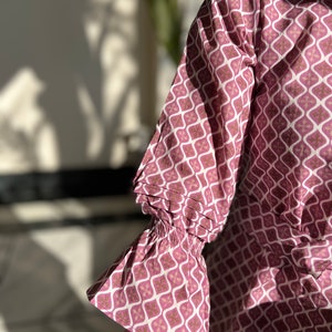 May include: A pink and white patterned blouse with a ruffled sleeve. The fabric has a geometric design.