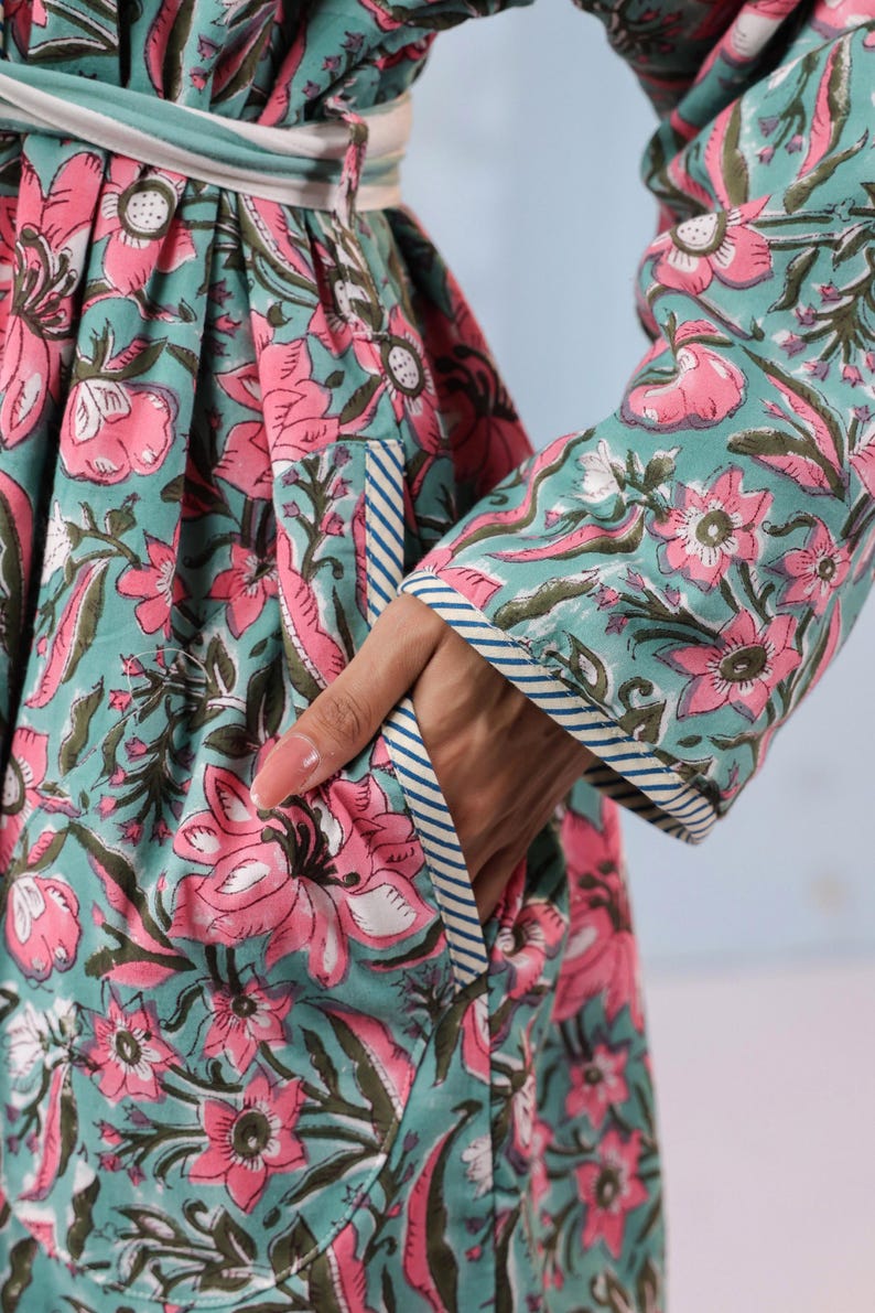 May include: A floral print robe in teal and pink. The robe features a large floral pattern with pink flowers and green leaves. The sleeves and edges are trimmed with a blue and white striped pattern. A hand is in the pocket.