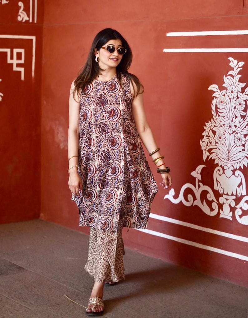 May include: A sleeveless tunic and matching pants set with a paisley pattern in shades of red, blue, and cream. The outfit is worn with sandals and sunglasses. The background is a red wall with white decorative elements.