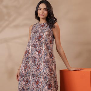 May include: Sleeveless, knee-length dress with a paisley pattern in red, blue, and cream. The dress has a high neckline and a loose fit. The model is wearing the dress and is leaning on an orange cube.