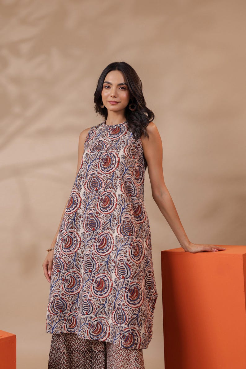 May include: Sleeveless, knee-length dress with a paisley pattern in red, blue, and cream. The dress has a high neckline and a loose fit. The model is wearing the dress and is leaning on an orange cube.