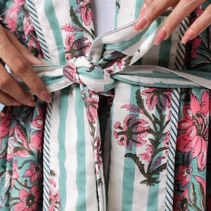 May include: A floral print robe with a striped pattern in white and teal. The robe features a tie closure and is adorned with pink and purple floral designs. The sleeves and edges are trimmed with a contrasting blue and white stripe.