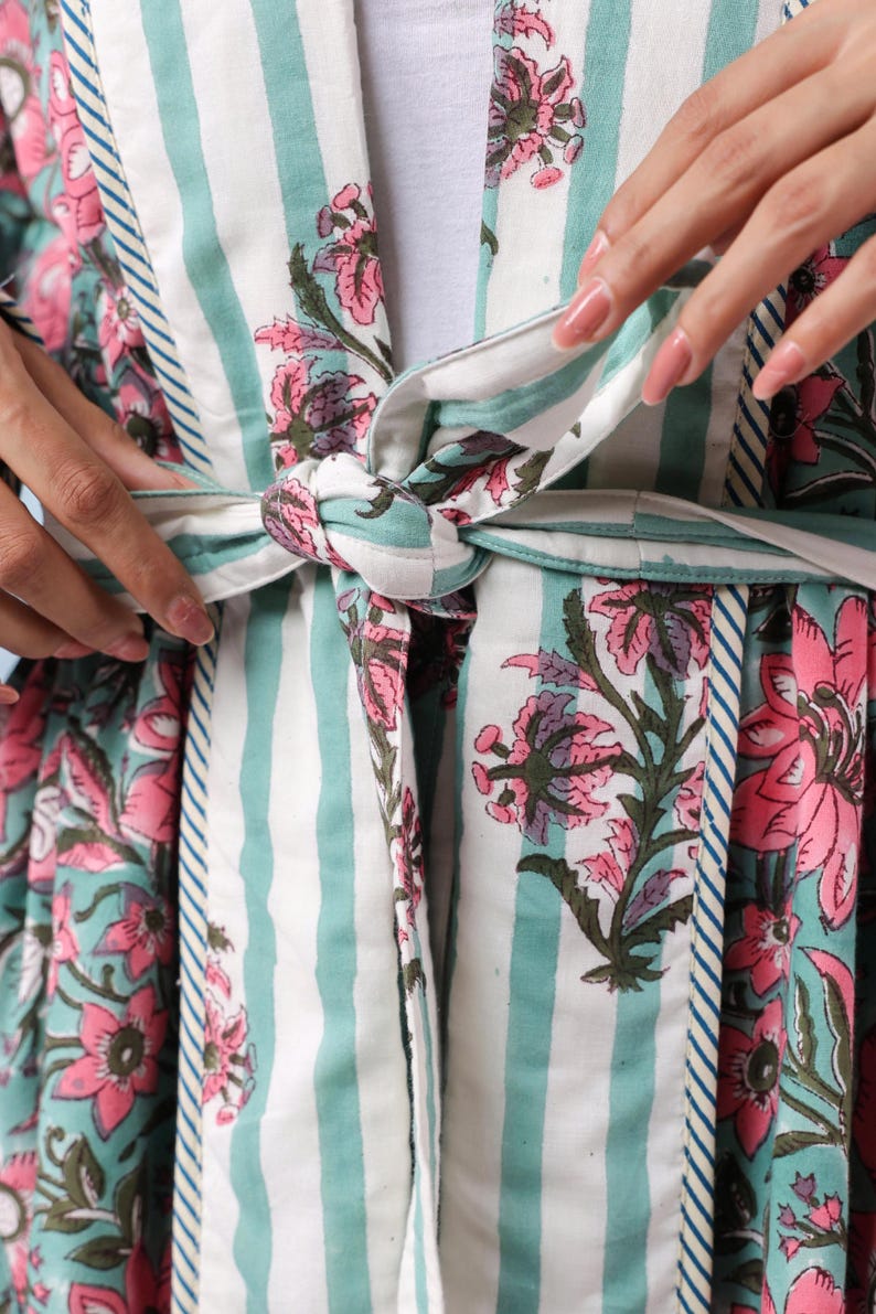 May include: A floral print robe with a striped pattern in white and teal. The robe features a tie closure and is adorned with pink and purple floral designs. The sleeves and edges are trimmed with a contrasting blue and white stripe.