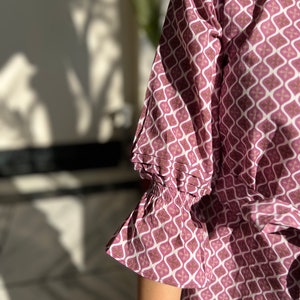 May include: A pink and white patterned blouse with a ruffled sleeve. The blouse has a geometric pattern with a white background and pink and brown accents.