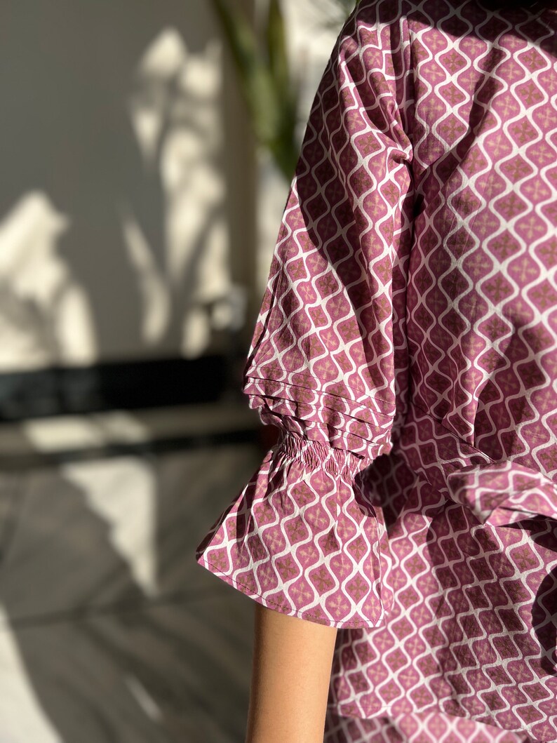 May include: A pink and white patterned blouse with a ruffled sleeve. The blouse has a geometric pattern with a white background and pink and brown accents.