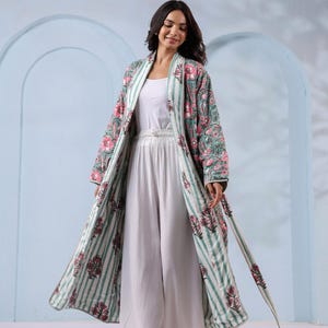 May include: A long, open-front robe with a floral pattern in pink and green, over a white outfit. The robe has a striped pattern on the inside and a matching belt. The outfit includes a white top and wide-leg pants. The background is a light blue.