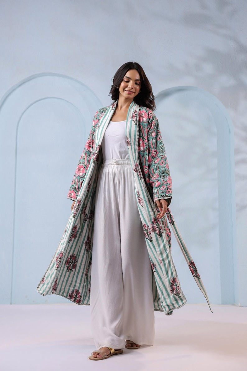 May include: A long, open-front robe with a floral pattern in pink and green, over a white outfit. The robe has a striped pattern on the inside and a matching belt. The outfit includes a white top and wide-leg pants. The background is a light blue.