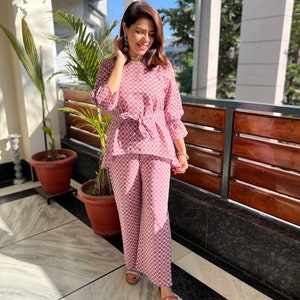 May include: A pink and white patterned pantsuit with a tie waist. The pantsuit is made of a lightweight fabric and has a relaxed fit. The model is wearing the pantsuit with sandals.