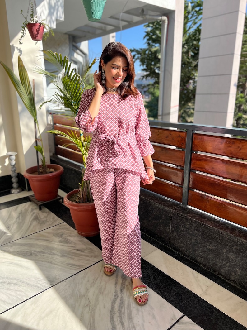 May include: A pink and white patterned pantsuit with a tie waist. The pantsuit is made of a lightweight fabric and has a relaxed fit. The model is wearing the pantsuit with sandals.