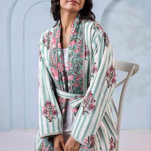 May include: A white and teal striped robe with a floral pattern on the collar and cuffs. The robe has a matching belt and is worn over a white outfit. The floral pattern features pink flowers and green leaves.