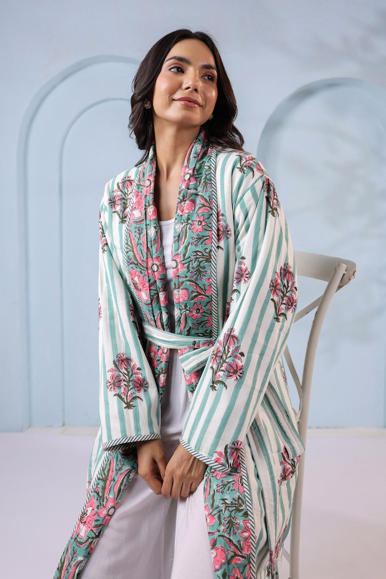 May include: A white and teal striped robe with a floral pattern on the collar and cuffs. The robe has a matching belt and is worn over a white outfit. The floral pattern features pink flowers and green leaves.